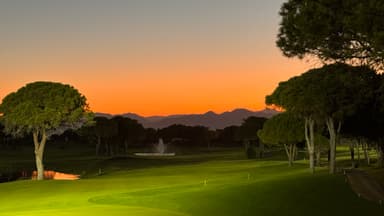 A serene golf course at sunset with trees and mountains in the background.