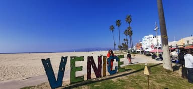 Une plage avec un grand panneau 'VENICE' et des palmiers sous un ciel bleu.