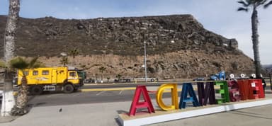 A colorful sign with the word 'TECATE' in front of a rocky hillside and a yellow truck on the road.