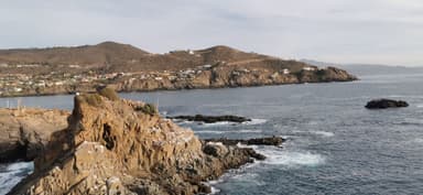 Rocky coastline with waves and distant hills under a clear sky.