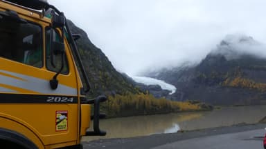 Un camion jaune près d'un glacier et des montagnes sous un ciel nuageux.