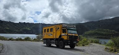 Un camion jaune sur une route près d'un lac entouré de collines verdoyantes.