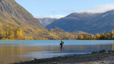 Un homme pêche dans un lac entouré de montagnes et d'arbres aux couleurs d'automne.