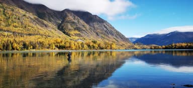 Un homme se tient dans un lac entouré de montagnes et d'arbres aux couleurs d'automne.