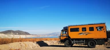 Un camion orange près d'un lac avec des montagnes en arrière-plan sous un ciel bleu.