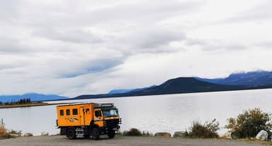 Un camion orange près d'un lac avec des montagnes en arrière-plan sous un ciel nuageux.