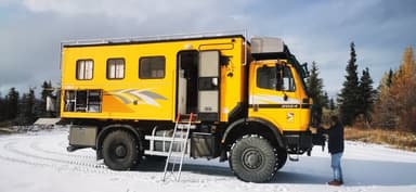 A bright yellow truck parked in a snowy landscape with trees in the background.