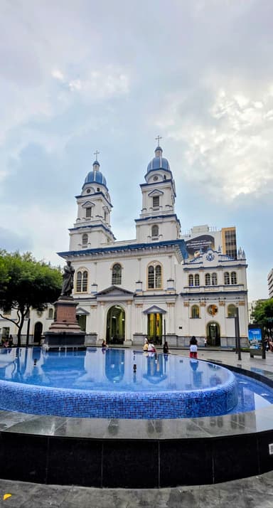 Une église blanche avec des tours et une fontaine devant, sous un ciel nuageux.