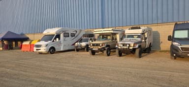 A row of various vehicles parked beside a blue building in the sunlight.