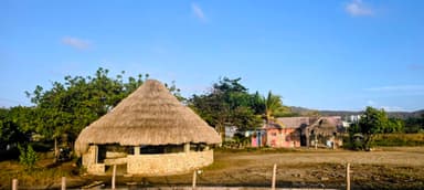 A thatched-roof hut beside colorful buildings under a clear blue sky.