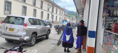 Deux personnes en vêtements traditionnels sur une rue de Silvia, Colombie, avec des voitures et des bâtiments en arrière-plan.