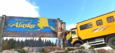Deux personnes posent devant un panneau 'Bienvenue en Alaska' sous un ciel bleu.
