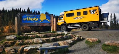 Un camion jaune près d'un panneau 'Bienvenue en Alaska' sous un ciel bleu.
