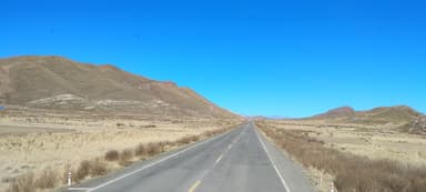 A long road stretches through a dry landscape with mountains under a clear blue sky.