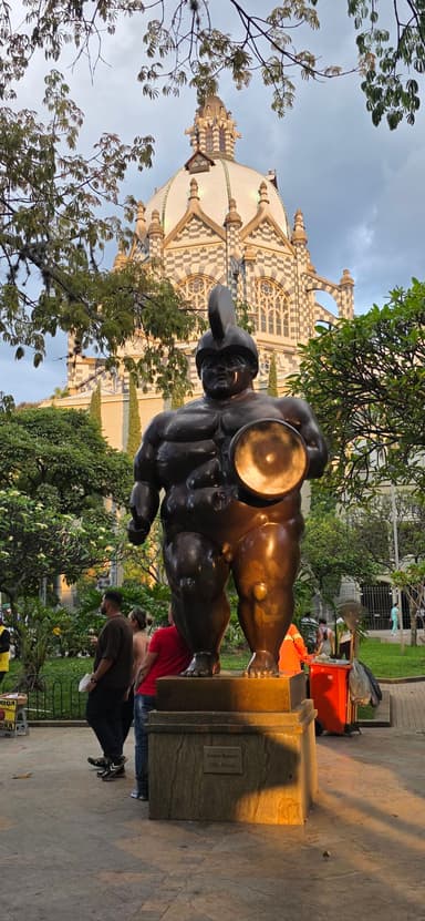 Une statue en bronze dans un parc avec un bâtiment en arrière-plan sous un ciel nuageux.