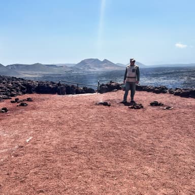 A person stands on red volcanic soil with mountains in the background under a clear blue sky.