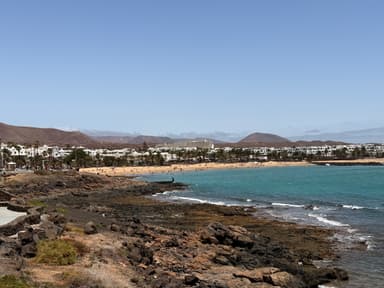 A scenic view of a beach with rocky shores and distant mountains under a clear blue sky.