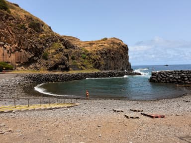 A rocky beach with calm water and people swimming under a clear sky.